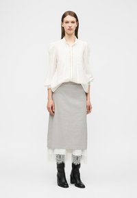 Γυναίκα όρθια, φοράει λευκό μπλούζα με μακριά μανίκια, γκρι φούστα μέσης μήκους με δαντελωτή όψη στο τελείωμα και μαύρα μποτάκια με τακούνι, σε μονόχρωμο φόντο.