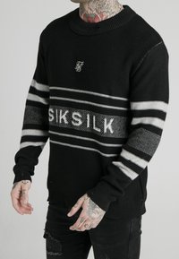 Svart stickad tröja med grå ränder och "SIKSILK"-logotyp. Ribbad krage och manschetter, med ett strukturerat mönster på ärmarna.