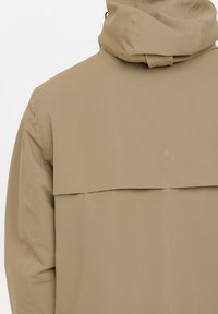 Beige Außenjacke mit strukturierter Oberfläche, mit hohem Kragen und einem Klappen-Design auf der Rückseite. Subtiles Logo-Detail in der oberen Mitte.