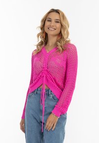 Rosa gehäkelter Pullover mit V-Ausschnitt, langen Ärmeln und Kordelzugdetail in der Taille, kombiniert mit hellblauen hochgeschnittenen Jeans.