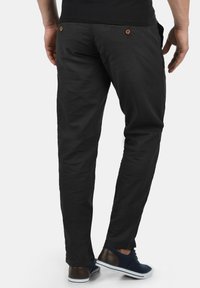 Pantalones negros de algodón con corte ajustado. Presentan dos bolsillos traseros con botones marrones y una textura suave, combinados con zapatos casuales.