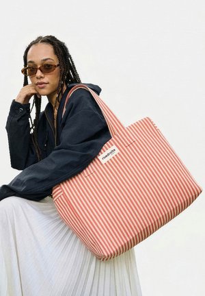 Femme aux tresses portant des lunettes de soleil marron et une veste sombre tenant un grand sac fourre-tout rayé rouge et blanc avec l'étiquette « rivedroite ».