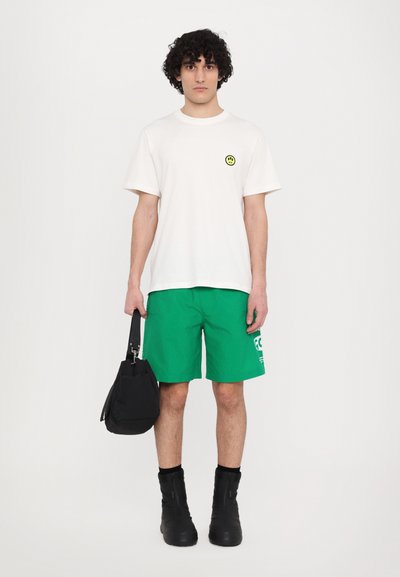 White t-shirt with a yellow smiley face graphic, green shorts with white print, black bag, and tall, black zip-up boots.