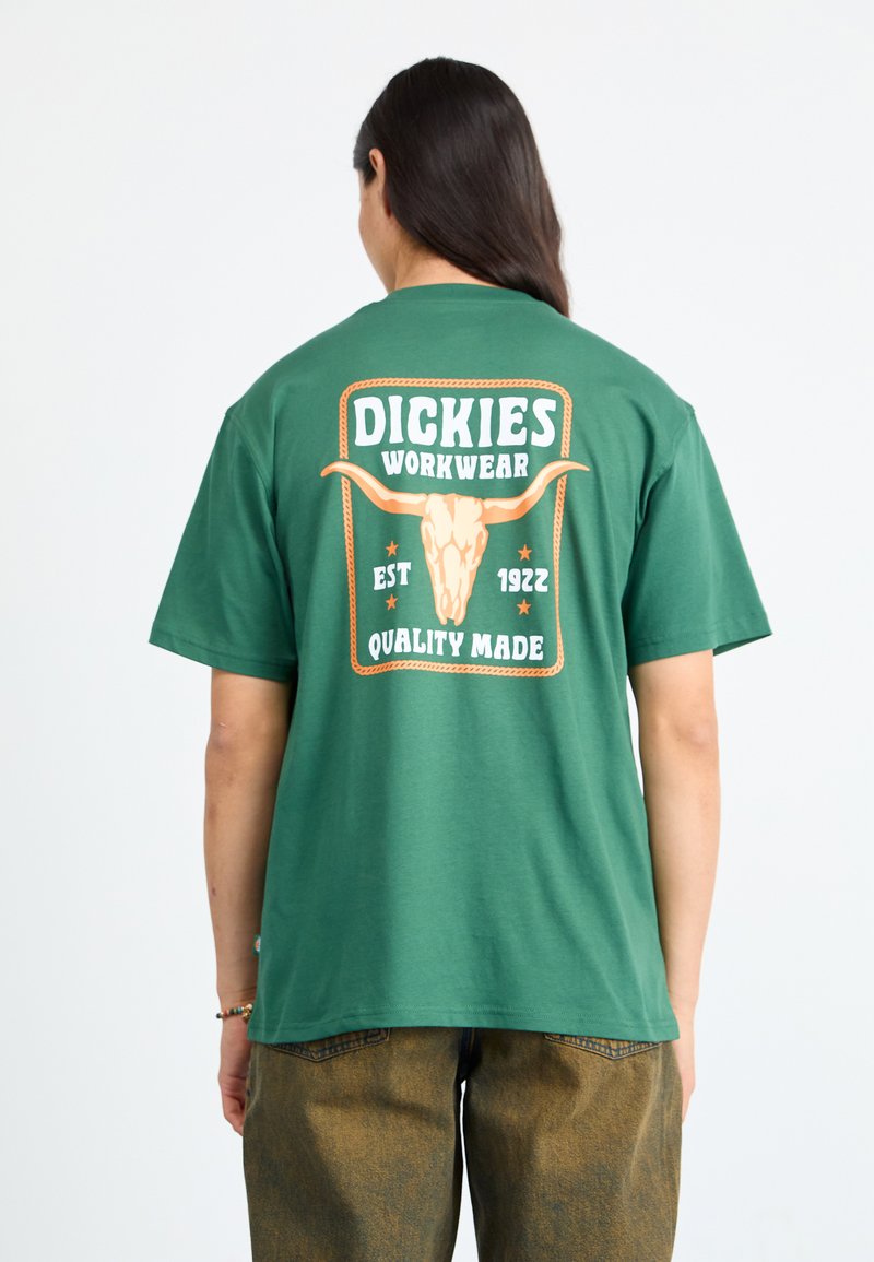 Zelena pamučna majica s grafikom na leđima koja prikazuje tekst "Dickies Workwear", lubanju bika i uzorke užeta, uz navedeni datum osnivanja i oznaku kvalitete.