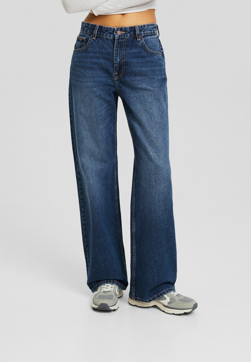 Weite blaue Jeans mit hohem Bund, fünf Taschen, kontrastierendem orangefarbenem Nähgarn und klassischem Knopfverschluss. Getragen mit grauen Sneakern.
