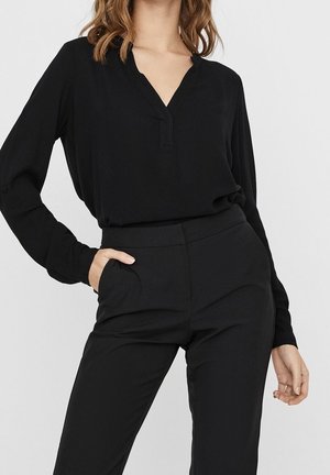 Vrouw draagt een zwarte blouse met lange mouwen, ingestopt in een zwarte broek met hoge taille, met een hand in de zak tegen een effen achtergrond.