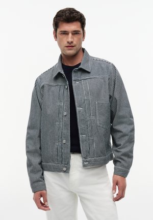 Mand iført sort og hvid vertikalt striped jakke over sort skjorte med hvide bukser, stående mod en ensfarvet hvid baggrund.