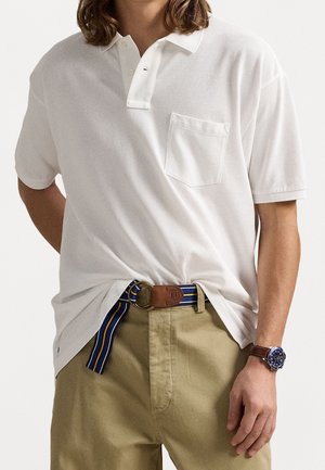 Biała koszulka polo z krótkim kołnierzykiem, pojedynczą kieszenią na piersi, w połączeniu z beżowymi spodniami i paskiem w paski. Na nadgarstku znajduje się zegarek.