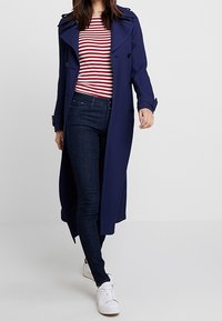 Kvinna som går klädd i en lång marinblå trenchcoat, randig röd och vit skjorta, mörka tajta jeans och vita sneakers.