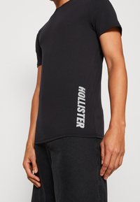 Černé tričko s krátkým rukávem vyrobené z lehkého materiálu, s vertikálním logem "HOLLISTER" v bílé barvě na levé straně.