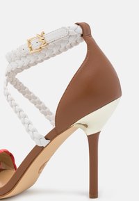 Sandale marron à talon haut avec un talon stiletto fin, bride de cheville tressée blanche et détail de boucle dorée.