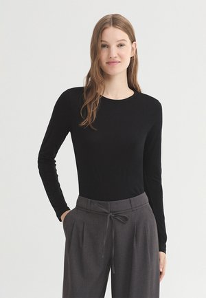 Junge Frau mit langen hellbraunen Haaren trägt ein eng anliegendes schwarzes Langarmshirt und lockere graue Hose mit gebundenem Gürtel, steht mit den Händen in den Taschen.