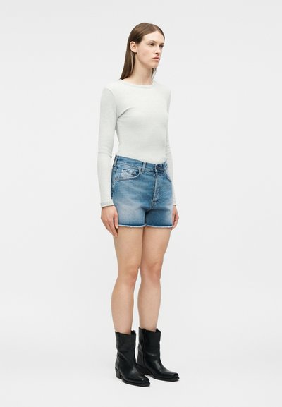 Γυναίκα που στέκεται φοράει εφαρμοστή λευκή μπλούζα με μακριά μανίκια, μπλε τζιν σορτς με ξεφτισμένα τελειώματα και μαύρα μποτάκια μέχρι τη μέση της γάμπας, σε λευκό φόντο.