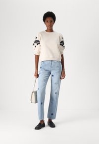 Lichtbeige sweatshirt met decoratieve zwarte bloemenborduursels, gecombineerd met lichtblauwe jeans met blauwe sterrenpatronen. Zwarte instappers en een grijze tas.