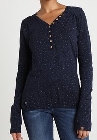 Mörkblå långärmad skjorta med små ljusblå och röda prickar, sju-knapp placket, bärs tillsammans med blå jeans.