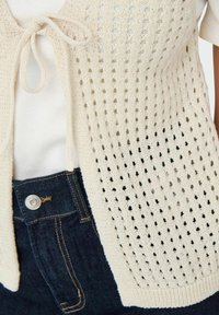 Light beige knitted cardigan with openwork design, tie closure, and short sleeves, paired with dark blue denim jeans featuring a buttoned waist.