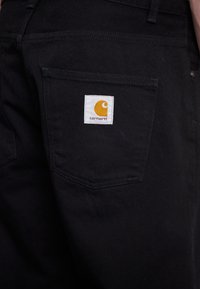 Detailní záběr na zadní kapsu černých kalhot s bílou a žlutou nášivkou loga Carhartt uprostřed kapsy.