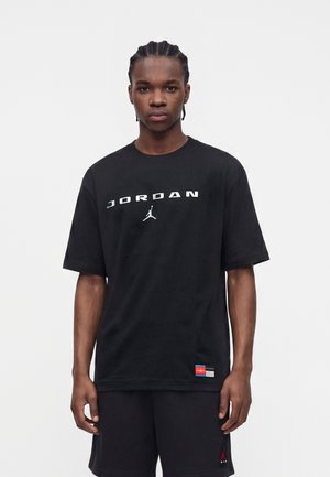 Junger Mann mit geflochtenem Haar trägt ein schwarzes Jordan-T-Shirt und schwarze Shorts und steht vor einem schlichten weißen Hintergrund.
