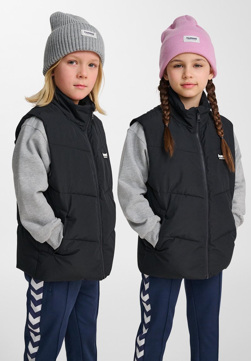 Zwei Kinder tragen ärmellose schwarze Steppwesten über grauen Langarmshirts, eines mit einer pinken Mütze und das andere mit einer grauen Mütze.