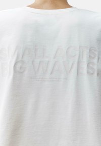 Balta medvilninė marškinėliai su laisvu pjūviu. Ant nugaros yra iškilūs užrašai "MAŽI VEIKSMAI, DIDELĖS BANGOS" su papildomais priežiūros nurodymais žemiau.