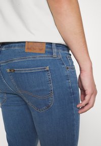 Vaqueros de mezclilla azul con una etiqueta de cuero en la cintura. Cuenta con bolsillos traseros con costuras visibles y un diseño ajustado.