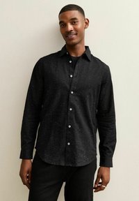 Zwarte button-up shirt met lange mouwen en een subtiel gespikkeld patroon. Voorzien van blauwe knopen en een klassieke kraag. Gedragen met zwarte broeken.