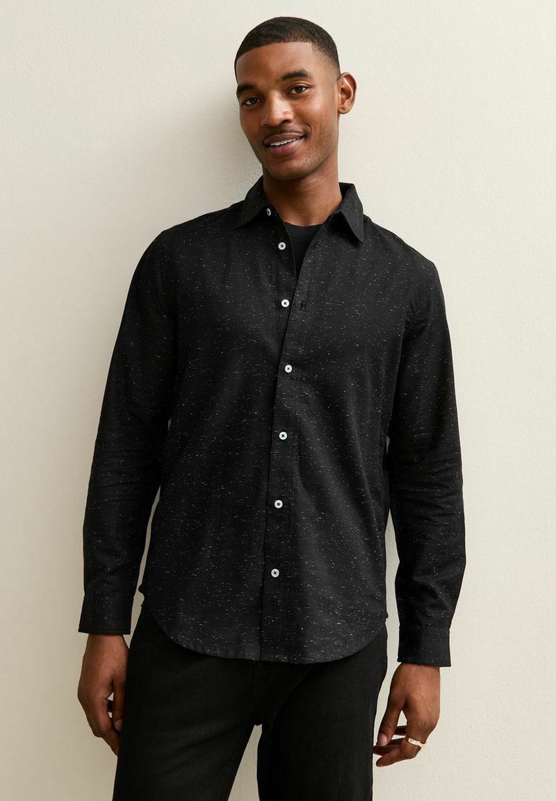 Zwarte button-up shirt met lange mouwen en een subtiel gespikkeld patroon. Voorzien van blauwe knopen en een klassieke kraag. Gedragen met zwarte broeken.