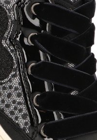 Zapatilla negra con cordones de terciopelo, ojales de charol brillantes y un panel lateral texturizado brillante con patrón de lunares.