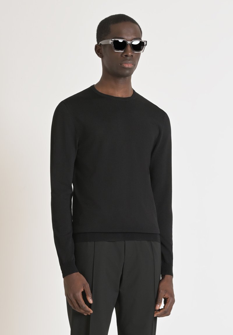 Maglione nero a maglia con collo rotondo, maniche lunghe e polsini a coste, abbinato a occhiali da sole neri. Ambientato su uno sfondo neutro.