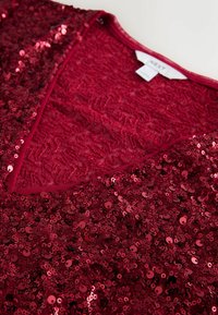 Haut en sequins rouges avec une surface texturée et un décolleté en V, orné de sequins circulaires brillants, avec une  étiquette de la marque NEXT.