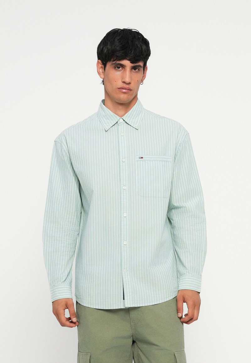 Chemise boutonnée rayée vert clair avec col, manches longues, poche poitrine unique et petit logo sur la poche. Tissu en coton.
