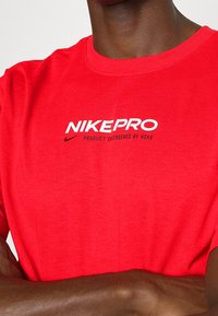 Punainen Nike Pro t-paita, lyhyet hihat, pyöreä kaula-aukko, puuvillasekoitekangas, jossa on valkoinen logosanoma "NIKEPRO" ja "TUOTEENTSUUNNITTELU NIKELTÄ".