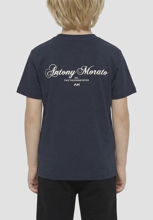 Blondes Kind trägt ein marineblaues T-Shirt mit dem Text „Antony Morato EST. TWO THOUSAND SEVEN AM“ auf dem Rücken, kombiniert mit schwarzen Hosen.