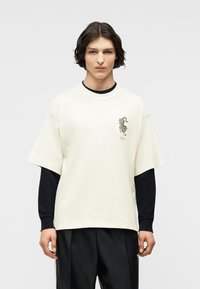 Νέος άνδρας με μεσαίου μήκους σκούρα μαλλιά που φοράει λευκό υπερμεγέθες T-shirt με μικρό γραφικό τί γρης πάνω από μαύρο μπλουζάκι με μακριά μανίκια.