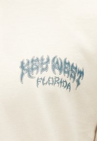 Μπεζ ύφασμα με μπλε στυλιζαρισμένο κείμενο που γράφει "HEY HEAST FLORIDA" σε γραμματοσειρά που μοιάζει με φλόγα, κεντραρισμένο στην επιφάνεια.