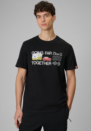 Młody mężczyzna w czarnej koszulce z grafiką czerwonego samochodu ciągnącego żółty przyczepę kempingową i napisem „Going Far Together” z przodu.