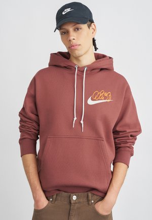 Sweat à capuche bordeaux avec des cordons blancs et un logo Nike stylisé orange sur la poitrine. Il possède une poche frontale et des poignets côtelés. Casquette noire portée.