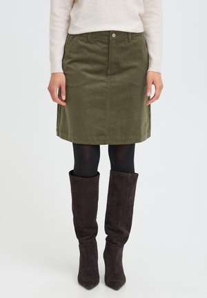 Femme portant une jupe en velours côtelé vert olive avec des collants noirs et des bottes mi-hautes en daim marron foncé, debout sur fond blanc