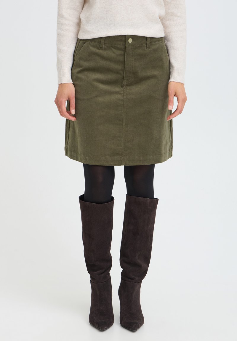 Femme portant une jupe en velours côtelé vert olive avec des collants noirs et des bottes mi-hautes en daim marron foncé, debout sur fond blanc