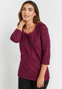 Top fucsia a maniche lunghe con motivo floreale nero, scollatura a barchetta e dettagli plissettati. Presenta bottoni sul davanti. Indossato con pantaloni neri.