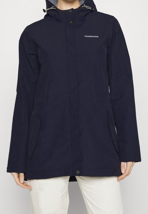 Persona che indossa una giacca a vento blu navy con cappuccio, tasche frontali e logo Didriksons sul petto, abbinata a pantaloni bianchi.