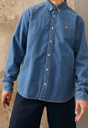 Personne portant une chemise en jean bleue à manches longues boutonnée avec un petit logo sur la poitrine, associée à un pantalon foncé.
