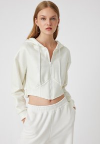 Felpa bianca cropped con zip e un leggero lucido, dotata di cappuccio con coulisse e tasche frontali. Abbinata a pantaloni jogger bianchi coordinati.