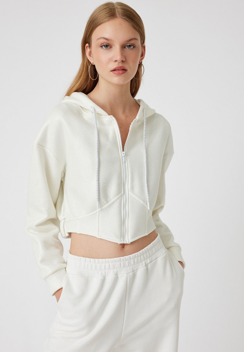 Felpa bianca cropped con zip e un leggero lucido, dotata di cappuccio con coulisse e tasche frontali. Abbinata a pantaloni jogger bianchi coordinati.