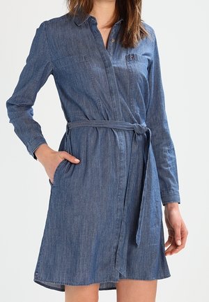 Femme portant une robe longue en jean bleu à manches longues avec ceinture à la taille et poches, debout devant un fond clair uni.
