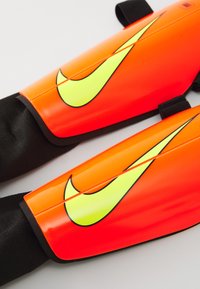Nike säärekatteid erksas oranžis koos peegeldava kollase logoga. Sile, vastupidav plastmaterjal mustade kangast rihmadega kindlustamiseks.