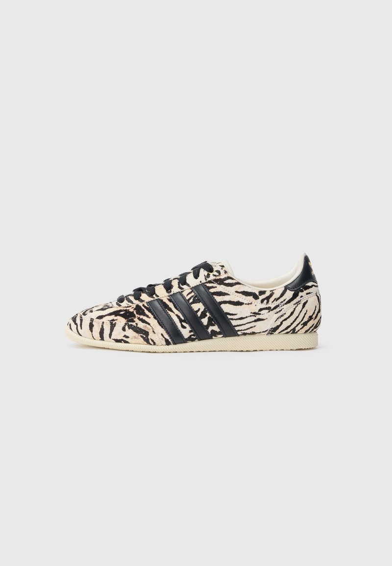 Adidas-sneakers med tigerstripemønster, med en beige og svart pelslignende tekstur, på en kremfarget gummisåle med svarte lisser og striper.