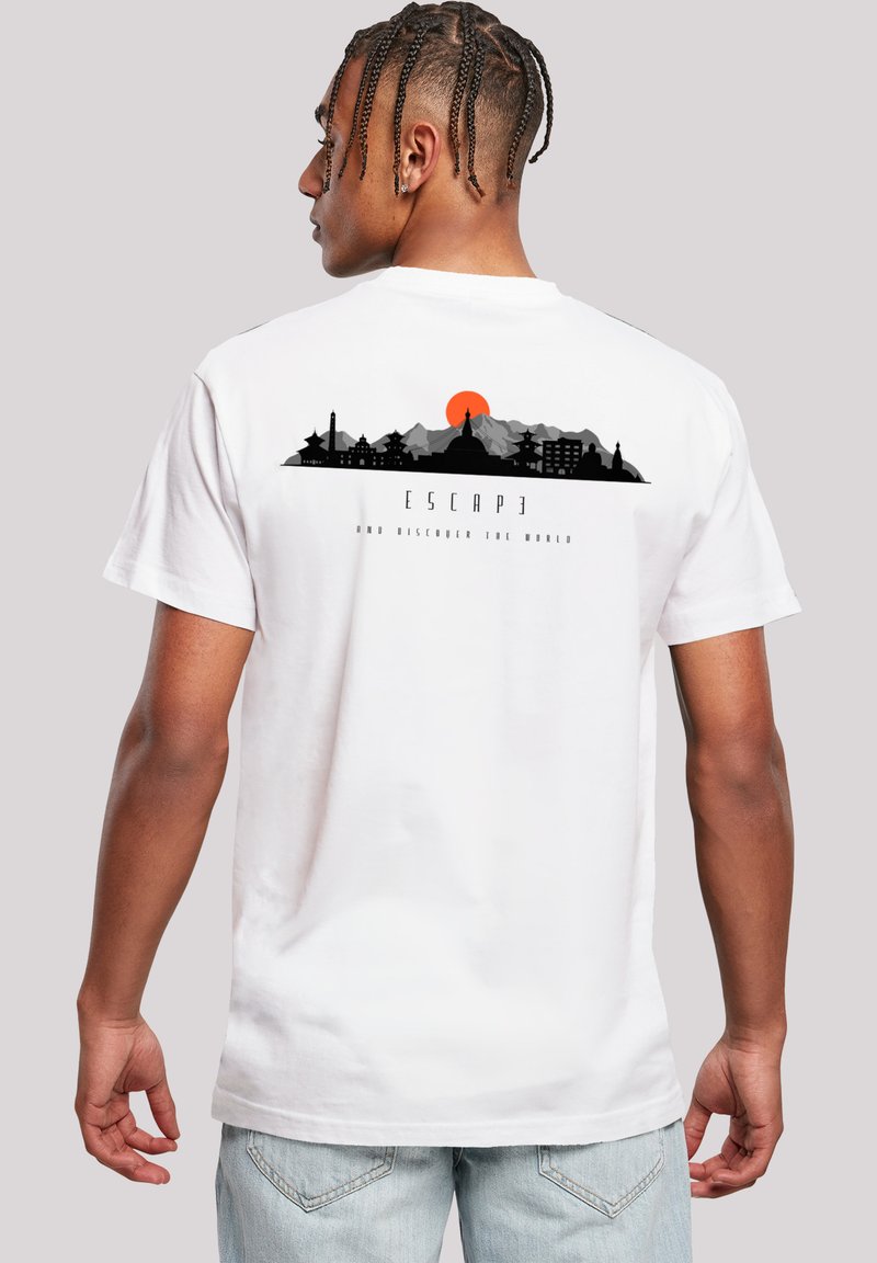 Mann mit geflochtenem Haar, der ein weißes T-Shirt trägt, auf dem eine schwarze Stadtsilhouette und eine rote Sonne darüber zu sehen sind, mit dem Text "ENTFLIEHEN und die Welt entdecken".