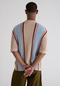 Knit short-sleeve shirt with beige, blue, and red vertical stripes. Textured fabric, ribbed collar, and relaxed fit. Worn with olive green pants.