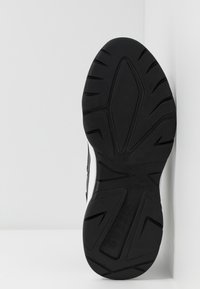 Semelle en caoutchouc noir d'une basket inclinée contre un coin blanc, montrant le motif de la semelle et le nom de la marque "GUESS" embossé sur la semelle.